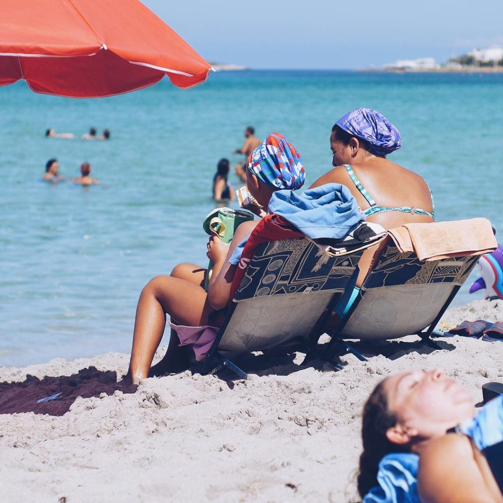 two ladies in hats sitting on sunloungers on the beach in Sicily facing blue sea 