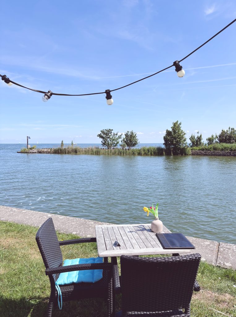 Rattan chairs and a wooden table with a tulip in a vase on the waterfront terrace at Paviljoen Strandbad Edam, with the Markermeer stretching out under a bright blue sky, Edam, North Holland