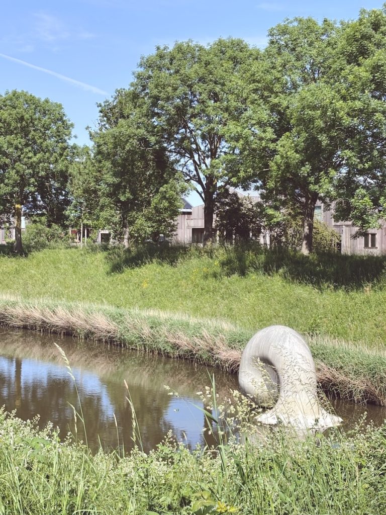 Abstract sculpture emerging from a canal beside wooden eco buildings surrounded by lush greenery, Vijfhuizen, North Holland