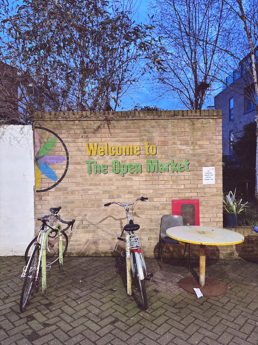 Welcome to The Open Market sign on a brick wall at Brighton Open Market, with bicycles parked outside