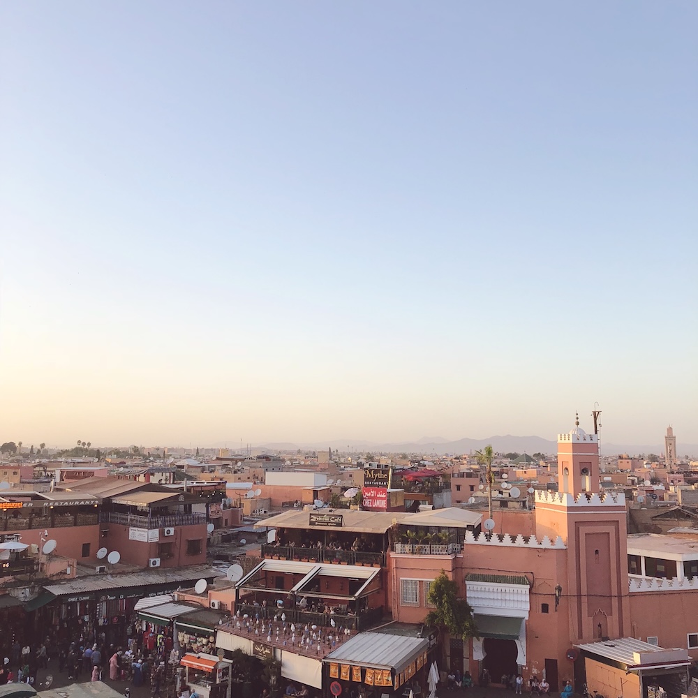 Jemaa el-Fna square in Marrakech at sunset, with rooftop restaurants, a pink minaret and the Atlas Mountains on the horizon