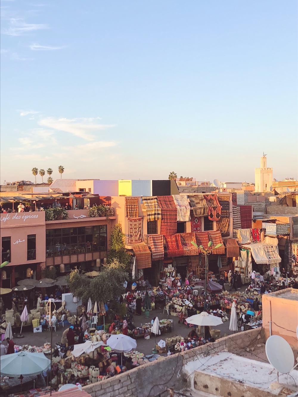 View over Rahba Kedima spice square in Marrakech at sunset, with rugs hanging from walls, market stalls and Café des Épices